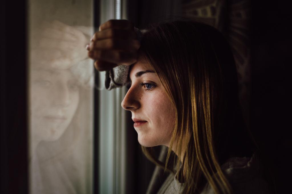 Femme regardant par une fenêtre vers un extérieur lumineux depuis un espace intérieur neutre, illustrant le désir de changement et la résistance à franchir le seuil vers l'inconnu.